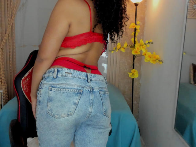 AriaVega with curly black hair, red lace bra, and blue denim jeans, showing her butt and red underwear, standing in front of a blue-covered chair and yellow flowers.