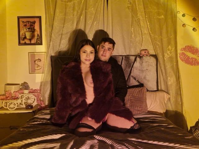 TinaYEdward with dark hair, wearing a purple fur coat, black lace stockings, and no pants, kneeling on a bed. TinaYEdward's ass is exposed, TinaYEdward's boyfriend sits behind her.
