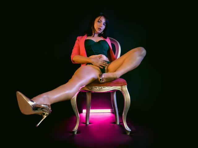 GrettaBorbon zittend op een gouden stoel met gespreide benen, gekleed in een roze jas, een groene top en zilveren hakken, die haar geslachtsdeel aanraakt, zwarte achtergrond, roze licht..