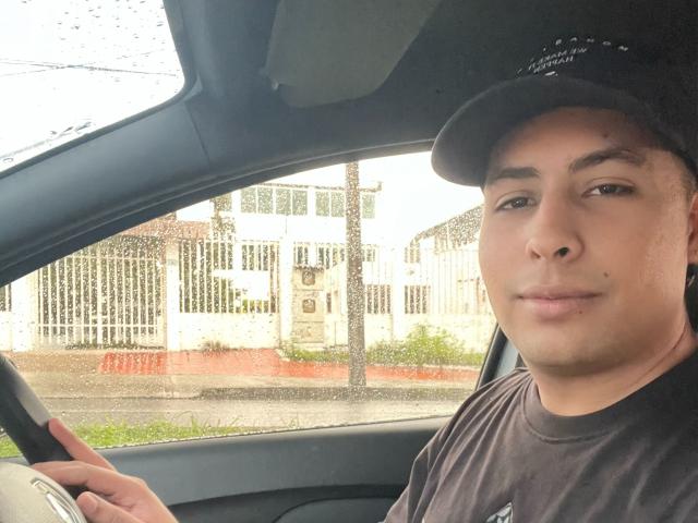 Sercho's face in a car, wearing a black cap, brown shirt, and a small mole on his left cheek. His hand is on the door, looking slightly bored.