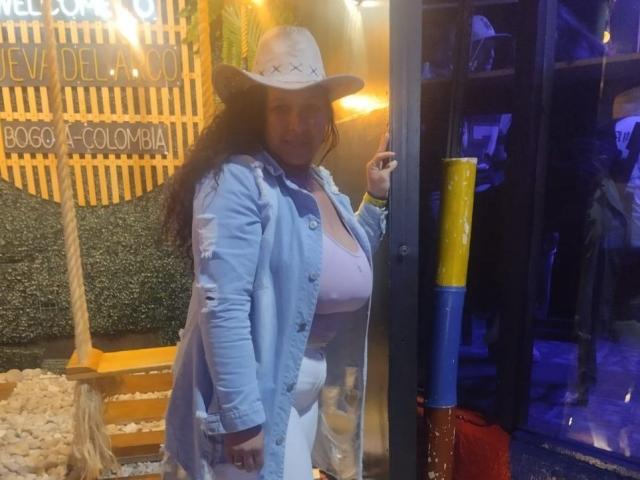 ZoehFerraro stands in a doorway, wearing a white cowboy hat, blue shirt, and white pants, showing off her large, round boobs, with a playful smile and hand on glass.