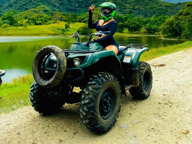 CloeEvanss with big ass, wearing black long-sleeve top, green helmet, and green visor, sitting on green ATV with big tires, touching helmet, on gravel road by lake.