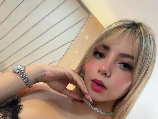 CloeEvanss with blonde hair, pink lipstick, and a diamond choker, wearing a silver watch, touching her cheek, showing her tits, and a black lace bra.