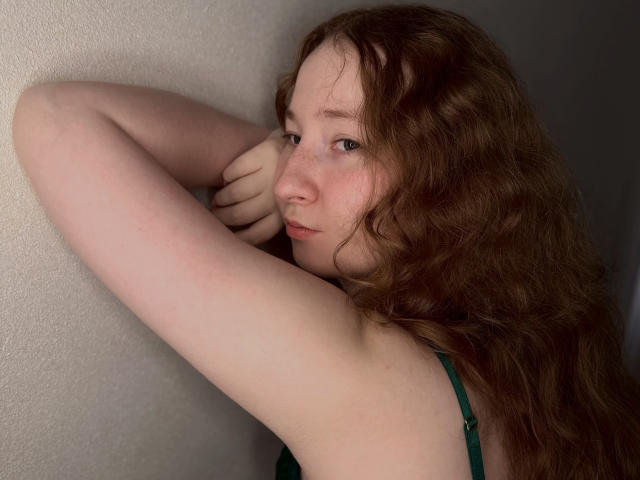 GeorgianneCoil with long, wavy brown hair, pale skin, and green eyes, leaning against a wall, arms raised, looking slightly to the side, wearing a green sleeveless top.