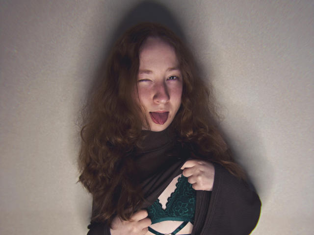 GeorgianneCoil with wavy red hair, black sheer top, green lace bra, tongue out, playful expression, hands on chest, pale skin, and slight blush.