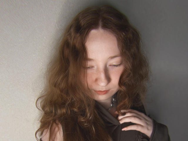 Georgianne, con lunghi capelli castani ondulati, occhi chiusi e un sorriso dolce, pelle chiara, tiene una camicia scura contro un muro grigio. La sua mano tocca il collo..