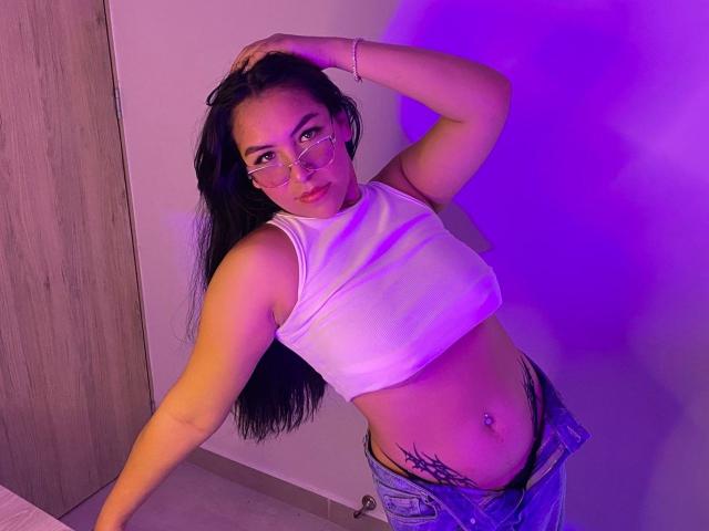 LiaDilucas with long black hair, wearing glasses, white crop top, and low jeans, showing a belly tattoo and a navel piercing, posing with hand in hair, purple light.