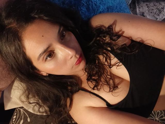 VelhMurphi, con cabello negro rizado, ojos marrones y labios rojos, vestida con una camiseta negra, mostrando sus pequeños pechos firmes, acostada sobre un cojín azul..