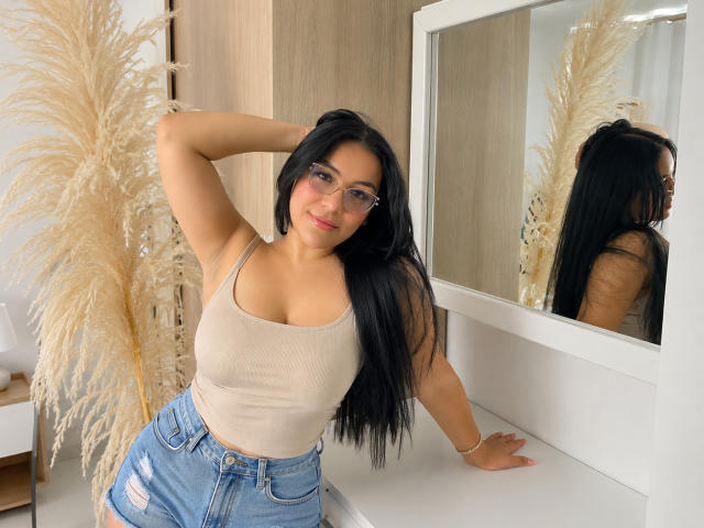 LiaDilucas with long black hair, glasses, beige tank top showing her tits, and blue jeans, leaning on a white counter, puffy beige pampas grass, mirror reflection.