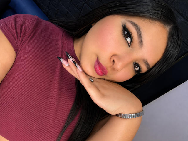 CharlotteTiffany with long black hair, wearing a maroon ribbed shirt, pouting pink lips, black eyeliner, and long black nails, touching her cheek with a silver bracelet on.