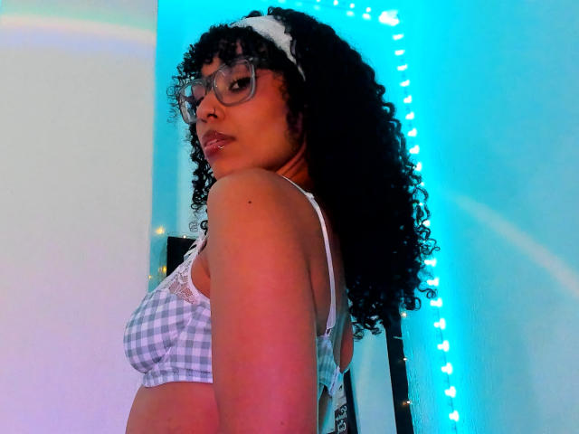 NickiBloom with curly black hair, wearing glasses and a white checkered bra, looking over her shoulder, her medium boobs visible, blue light in the background.