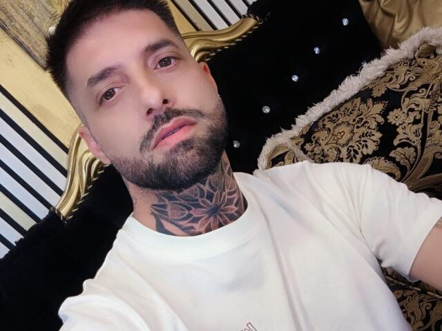 TommyDum with a tattooed neck, white shirt, short brown hair, beard, and brown eyes, sitting on a black and gold upholstered couch. His face is slightly tilted downward.