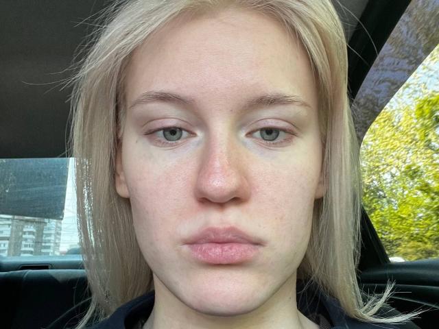 AnglaEntrekin a la peau pâle, les cheveux blonds et lisses, les yeux verts et les lèvres rosées, avec une expression neutre, et elle est assise dans une voiture. Son petit nez, légèrement retroussé, est visible..