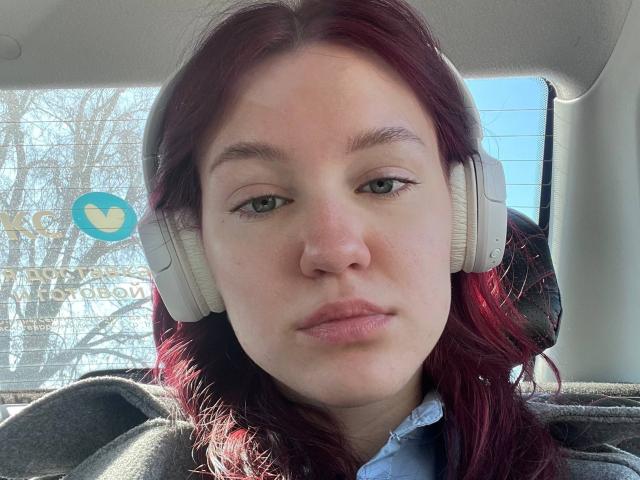 AnglaEntrekin, avec ses cheveux roux, ses yeux verts et ses lèvres roses, portant des écouteurs blancs, a une expression neutre et se trouve à l'intérieur d'une voiture baignée de lumière du soleil. Elle porte une chemise bleue..