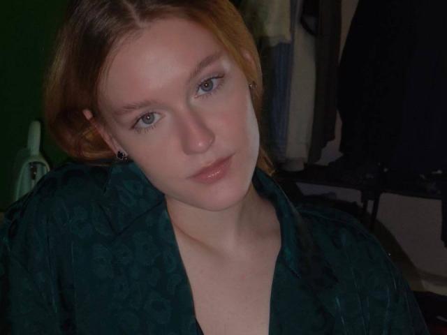 AnglaEntrekin a la peau pâle, les cheveux roux et les yeux bleus, elle porte une chemise verte à motifs, avec les lèvres légèrement entrouvertes et une expression neutre, dans une pièce faiblement éclairée..