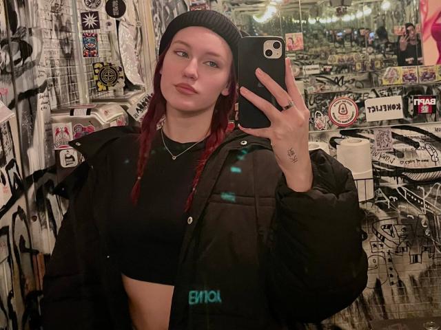 AnglaEntrekin, avec des tresses rouges, portant un bonnet noir, une veste matelassée et un crop top noir, prend un selfie devant un miroir avec un iPhone. Tatouage sur la main droite..