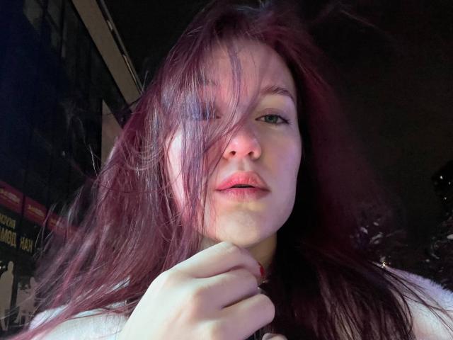 AnglaEntrekin, avec ses cheveux roux en bataille, sa peau claire et ses lèvres roses, se tient la menton, laissant entrevoir un maquillage à lèvres discret et un petit nez. Ses yeux verts sont légèrement tournés sur le côté..
