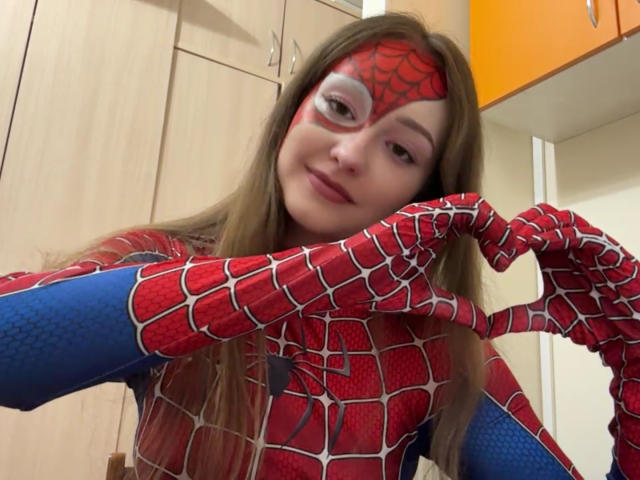 MinhMajic, avec ses longs cheveux bruns, son costume de Spider-Man rouge et bleu, son masque et ses gants à motifs de toile d'araignée, formant un cœur avec ses mains, souriant légèrement..