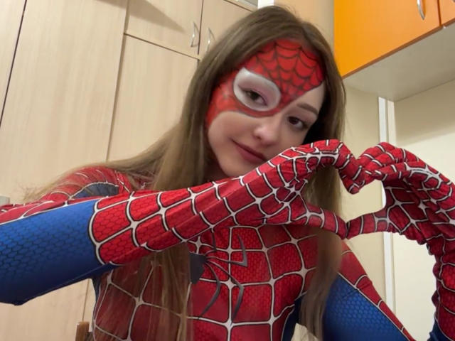 MinhMajic, avec de longs cheveux bruns, portant une combinaison rouge et bleue de Spider-Man, formant un cœur avec ses mains, souriant à la caméra..