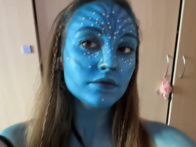 MinhMajic a du maquillage bleu sur le visage avec des points blancs, des cheveux bruns et une expression sérieuse. Ses épaules sont dénudées. L'image est un autoportrait avec un arrière-plan légèrement flou..