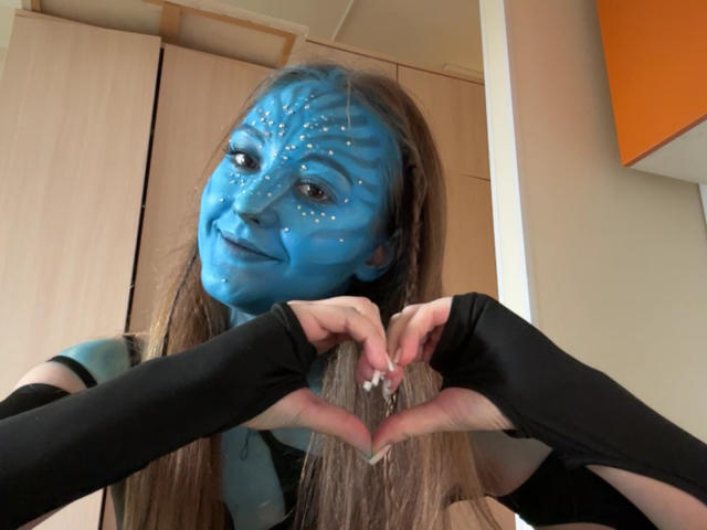 MinhMajic, avec du maquillage bleu sur le visage, des manchettes noires et de longs cheveux bruns, forme un cœur avec ses mains, regarde la caméra et a des paillettes blanches sur son visage..