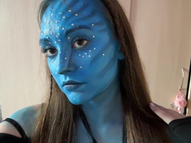 MinhMajic hat blaue Gesichtsbemalung mit weißen Tupfen, braune Augen und langes, braunes Haar. Ihre Hände sind sichtbar, eine hält ein pinkes Objekt. Sie trägt ein schwarzes Oberteil..