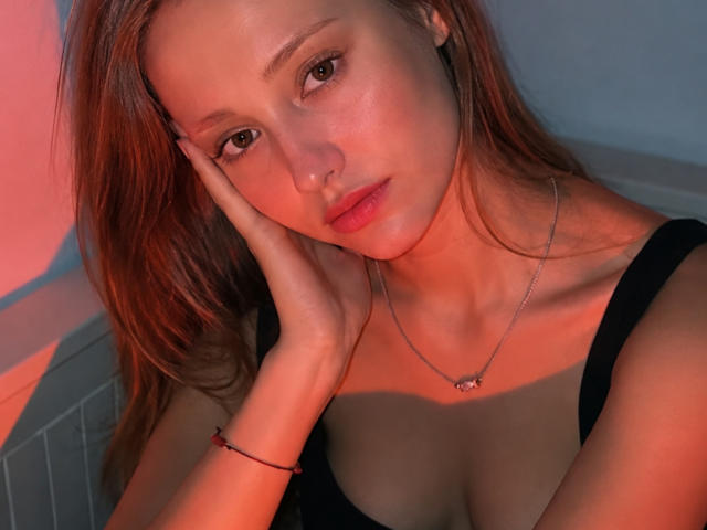 MinhMajic mit roten Haaren, Hand am Wangen, blickt in die Kamera, trägt ein schwarzes Oberteil, zeigt ihre Brüste, eine silberne Halskette, ein rotes Armband, weiches orange-blaues Licht, leichtes Schmollen, sanfter Ausdruck..