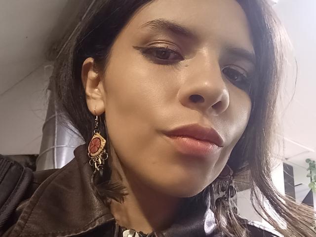 Das Gesicht von VioletaStone, braune Augen, Eyeliner im "Winged"-Stil, roter Lippenstift, leichtes Lächeln, braune Lederjacke, aufwendige Ohrringe in Rot und Schwarz, braunes Haar, weiche Haut, dezentes Make-up, leicht spitze Nase..