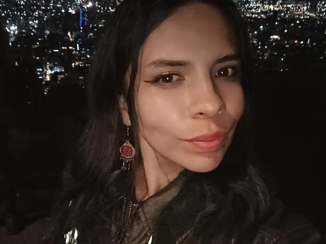 VioletaStone, avec ses cheveux foncés, ses yeux marrons et ses lèvres roses, porte un boucle d'oreille rouge orné d'une gemme, dans une nuit sombre illuminée par les lumières de la ville. Son nez est légèrement retroussé..