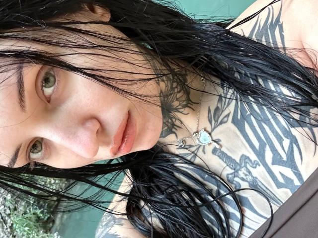 NyxelleSable has wet black hair, green eyes, and tattoos covering her chest and neck. She's looking at the camera with slightly parted lips. Her tits are small.