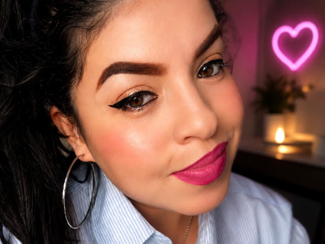 AlessiaLuxe has dark hair, brown eyes, pink lipstick, and black eyeliner. She wears a blue shirt and large hoop earrings. A pink neon heart is in the background.