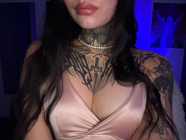 NyxelleSable with large boobs, tattooed neck and chest, wearing a pink satin dress, black choker, and long dark hair, has glossy pink lips and a knife tattoo on chest.