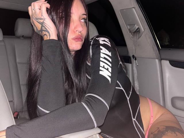 NyxelleSable with long black hair, nose ring, and tattoos, wearing a black long-sleeve top and pink thong, sitting in a car, looking to the side, hand in hair.