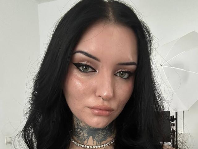 NyxelleSable with dark, wavy hair, green eyes, and black eyeliner, wearing a choker, has a tattoo on her neck, and a shiny, slightly flushed face.