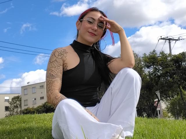 Amy, capelli rossi con punte colorate, occhiali, top nero senza maniche, pantaloni bianchi, braccio con tatuaggi, seduta sull'erba, mano sulla testa, cielo blu, nuvole bianche, sorridente, carina, sexy..