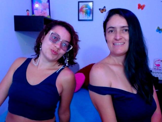 LunaVenus with short brown hair and glasses pouts, wearing a black crop top. LunaVenus with long black hair smiles, wearing a black off-shoulder top. Both have tits.