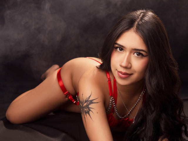 HannaThorne with long black hair, red lingerie, and a tattoo on her upper arm, lying on her stomach, smiling at the camera. Her ass is raised, looking sexy.