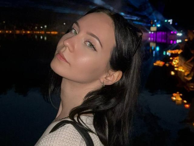Dennise Deschepper, con la pelle chiara, gli occhi verdi e i capelli neri, indossa una maglietta bianca, guarda oltre la spalla, sorridendo leggermente, in un contesto urbano notturno, scuro e suggestivo..