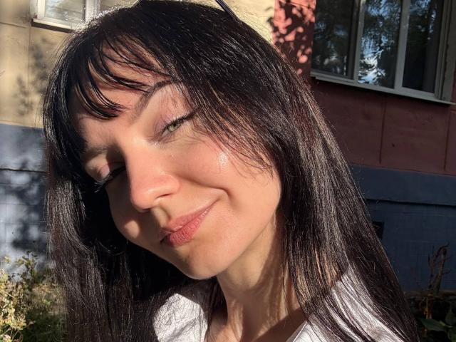 El rostro de Dennise Deschepper, con cabello negro y lacio, ojos verdes ligeramente entrecerrados, labios rosados claros y una sonrisa suave, iluminado por el sol y con sombras, mostrando pecas sutiles y una textura de piel delicada..