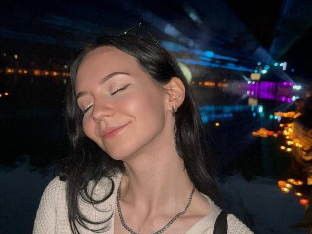 DenniseDeschepper with closed eyes, smiling, pale skin, black hair, white sweater, small earrings, necklace, dark night background with colorful lights reflecting on water.