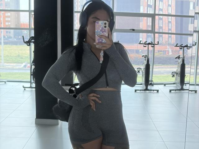StellaBeckker with headphones, gray zip-up crop top, and matching shorts, holding phone in mirror selfie, hand on hip, black bag, standing in gym with exercise bikes.