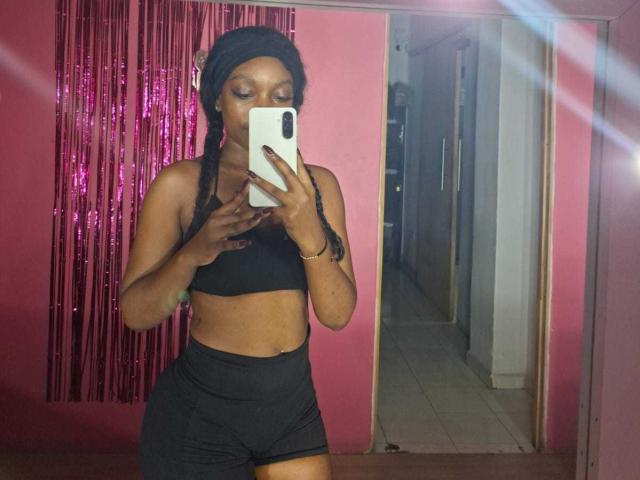 ColombianEbonyVibes with dark skin, black sports bra, black shorts, and headwrap taking a mirror selfie, holding a white phone, standing in a pink room with pink curtains.