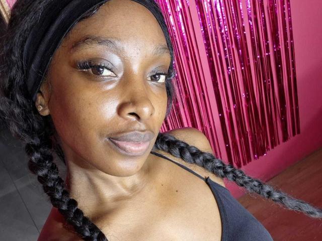 ColombianEbonyVibes ha la pelle scura, lunghi capelli neri intrecciati, indossa una canotta nera, con tende rosa lucide sullo sfondo. Le sue labbra sono leggermente dischiuse e ha un sorriso appena accennato..