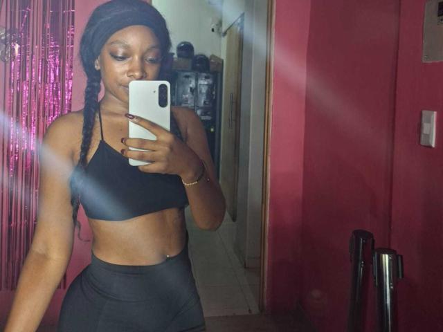 ColombianEbonyVibes prend un selfie devant un miroir, vêtue d'un soutien-gorge de sport noir et d'un pantalon noir taille haute, avec de longues tresses et un bonnet, tenant un téléphone blanc, montrant ses abdominaux bien dessinés..