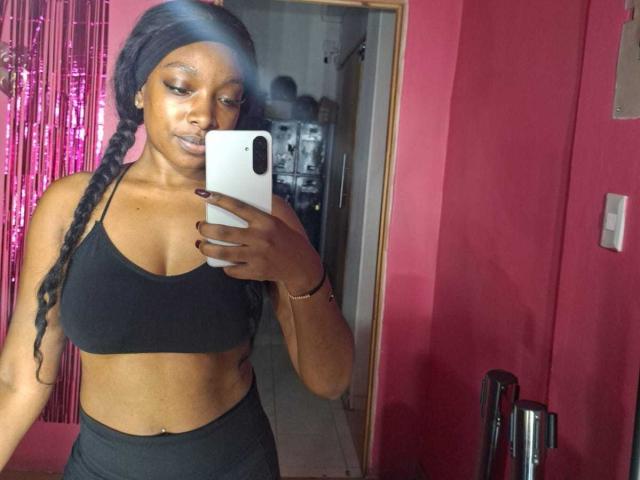 Atmosfera colombiana: una donna con la pelle scura, un reggiseno nero e pantaloni neri, che tiene in mano un telefono bianco, in una stanza rosa, con il suo riflesso in uno specchio, con lunghi capelli trecciati e un trucco leggero..
