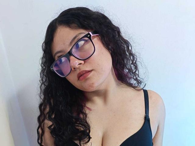 SarahThomsonn with curly black hair, wearing glasses and a black bra, showing her boobs, looking slightly down, with a neutral expression.