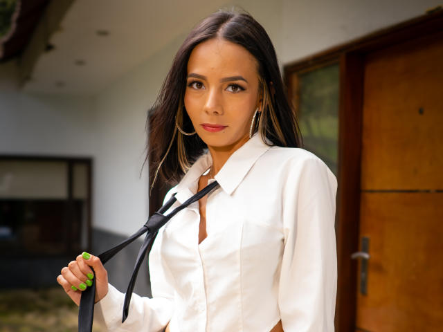 EvelinLuna with long dark hair, wearing a white button-up shirt, holding a black tie, and gold hoop earrings, standing in front of a wooden door. Her green nails.