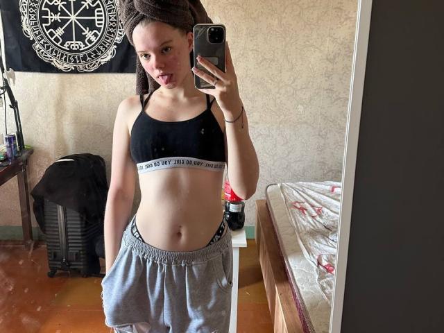 DominicaChambley with a towel on her head, black sports bra, and grey sweatpants, sticking out her tongue, taking a mirror selfie, showing her flat stomach and small tits.