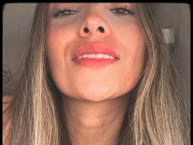 El rostro de AdharaCooperr, mostrando una ligera sonrisa con labios rosados, cabello castaño liso con reflejos rubios, y una pequeña peca negra en el lado izquierdo de su barbilla..