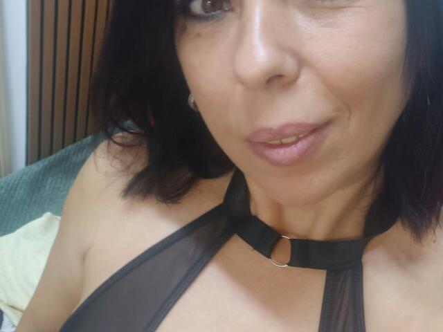 LauraSin, com cabelo escuro, piscando, vestindo uma blusa preta transparente com alças e um colar de metal, mostrando seus pequenos seios e lábios rosados. Close-up..
